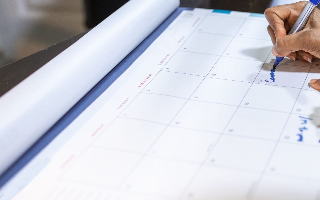 A hand marking dates on a paper desk calendar