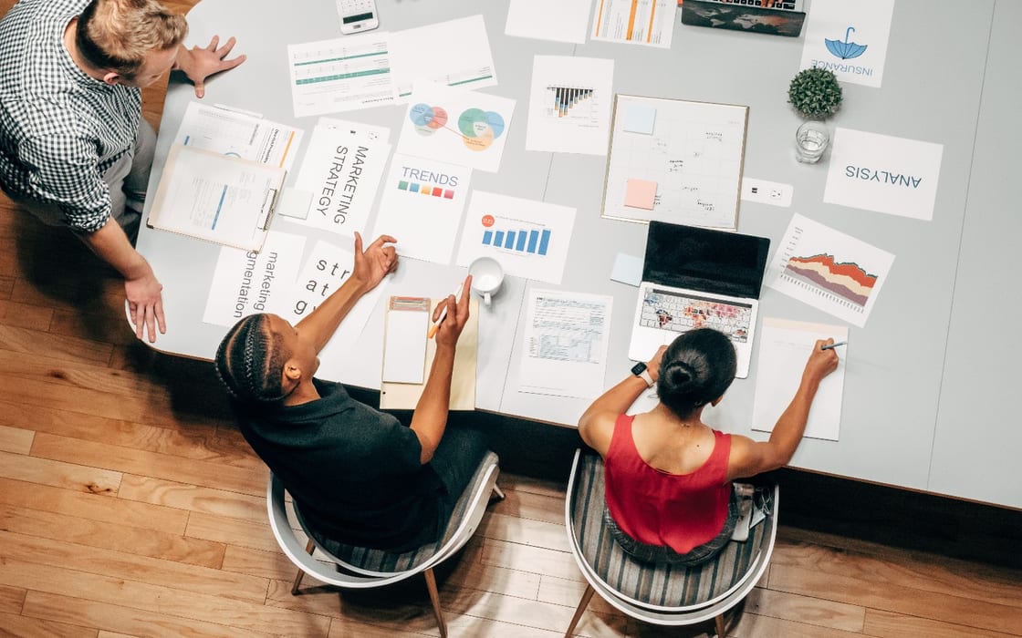 Three people around a table covered in printed marketing strategy, trends and analysis sheets, seen from above