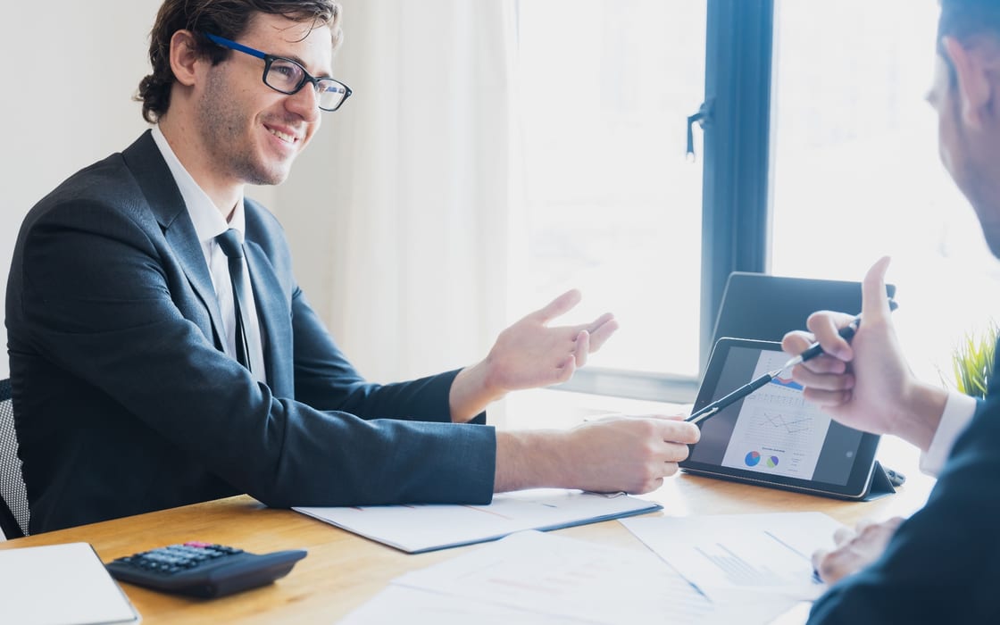 Two people in a meeting, one turning a tablet of charts towards the other across the desk